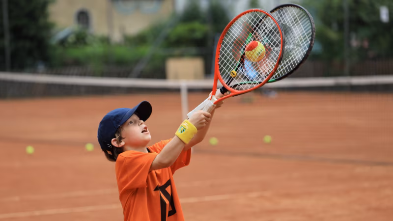 Junior catching a tennis ball using two tennis rackets on a clay court